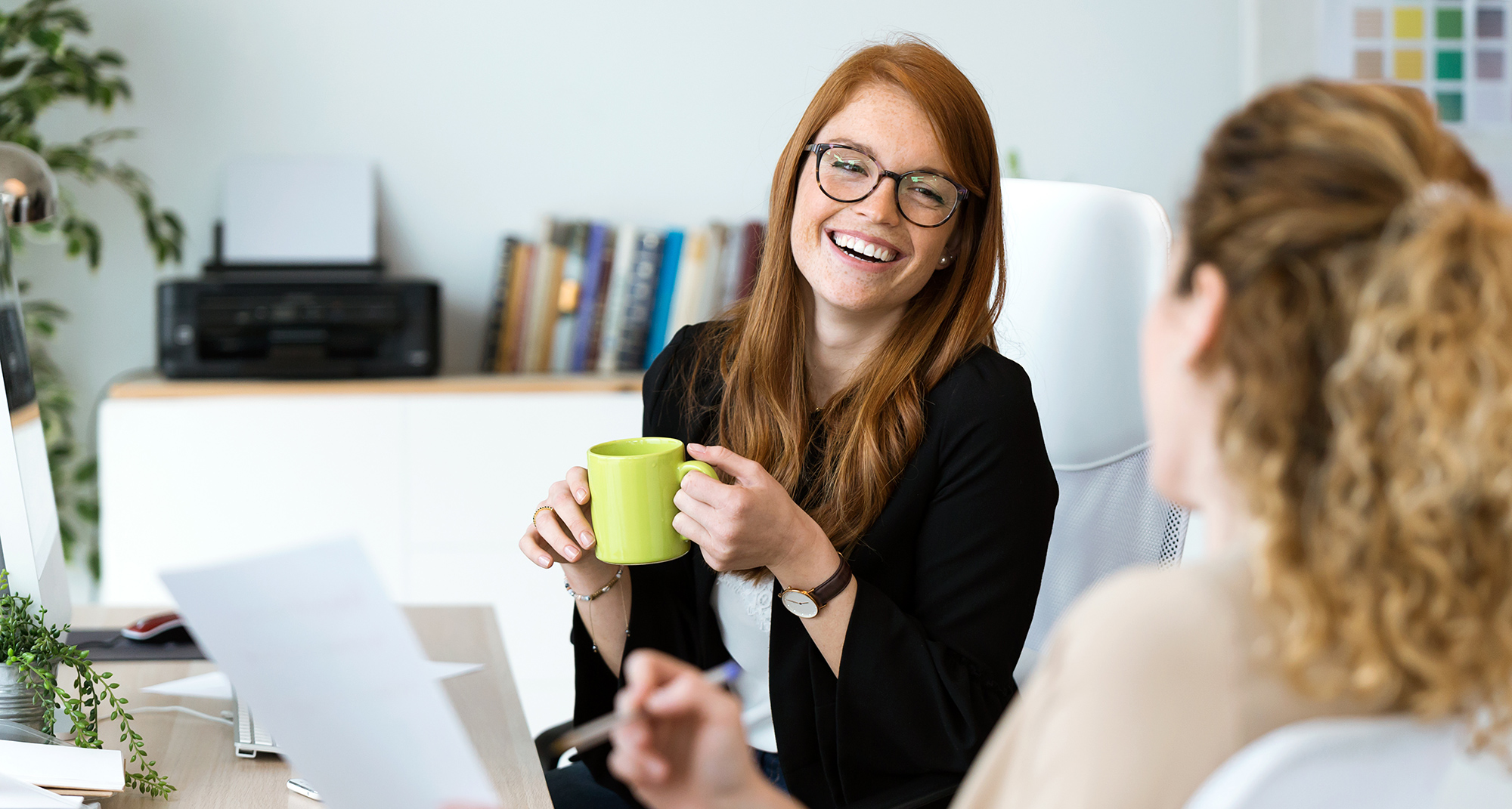 A woman with glasses holding a mug, laughing with her co-worker