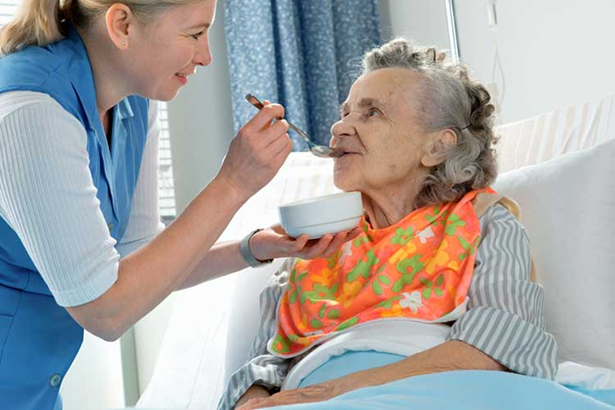 A support worker feeding an elderly woman in a hospital bed